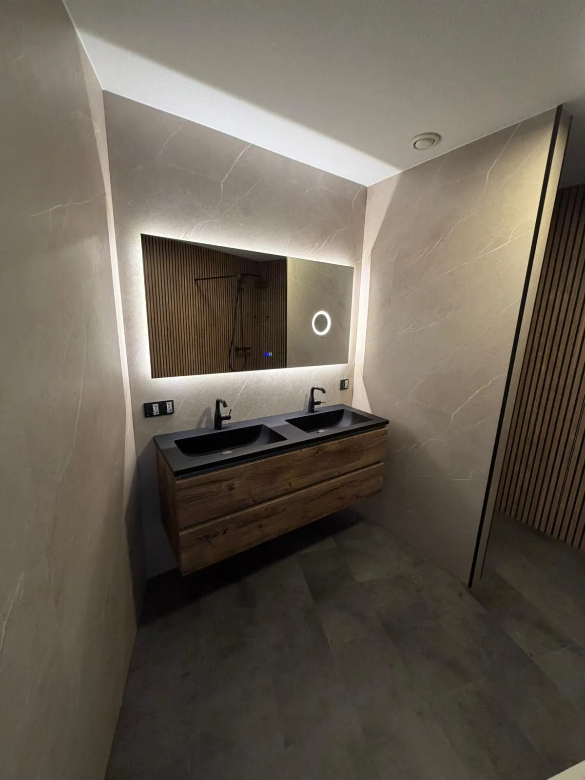 Een moderne badkamer met een dubbele wastafel van hout met zwarte keramische bassins en twee zwarte kranen. De muren zijn betegeld met grote, grijze marmerlook tegels. Er is een ingebouwd LED-licht boven het spiegelkastje.