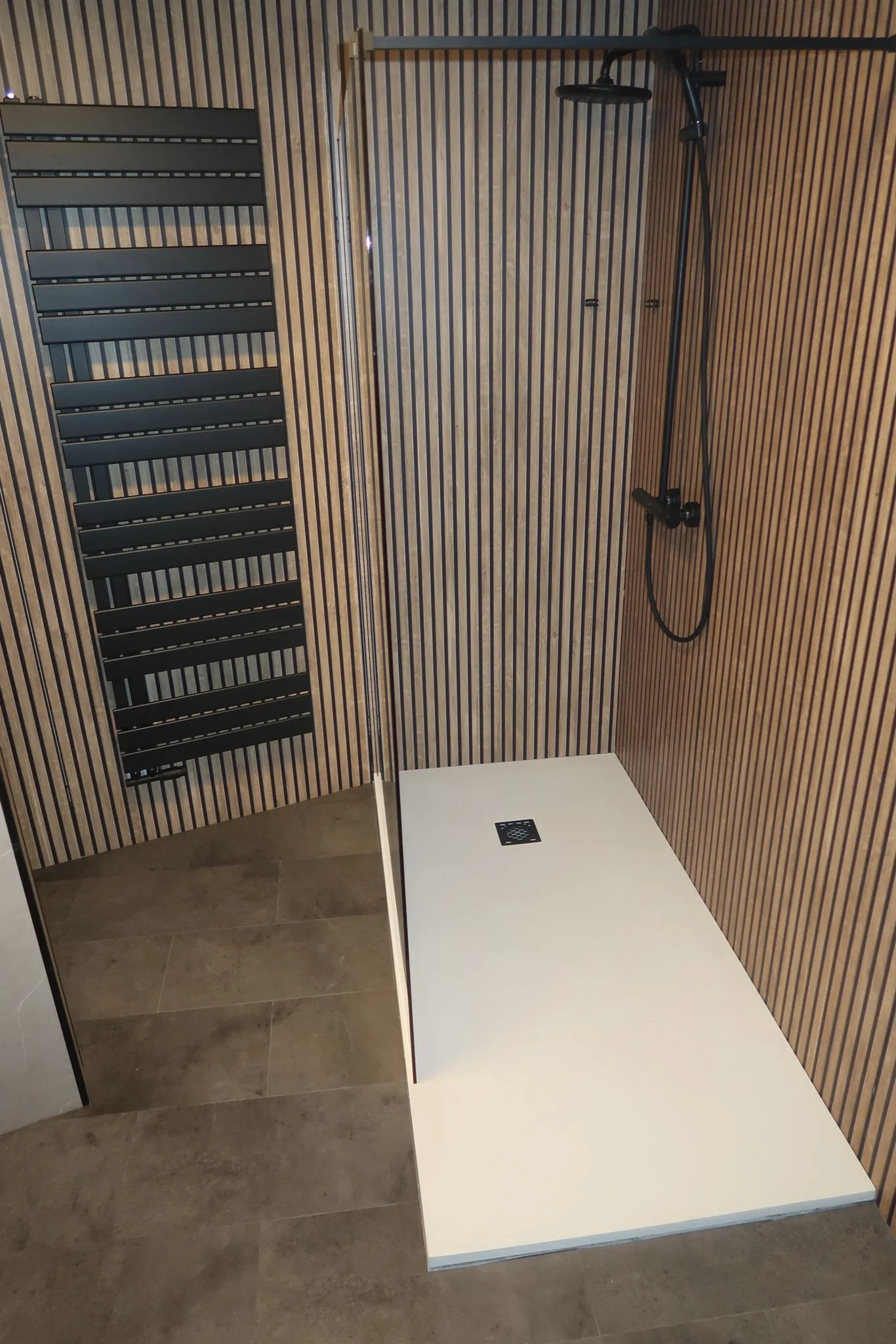 Een moderne douche met een witte douchebak, betegelde wand en vloer. De wand heeft verticale, bruine en zwarte strepen. Er is een zwarte douchekop en een verwarmingselement aan de wand.