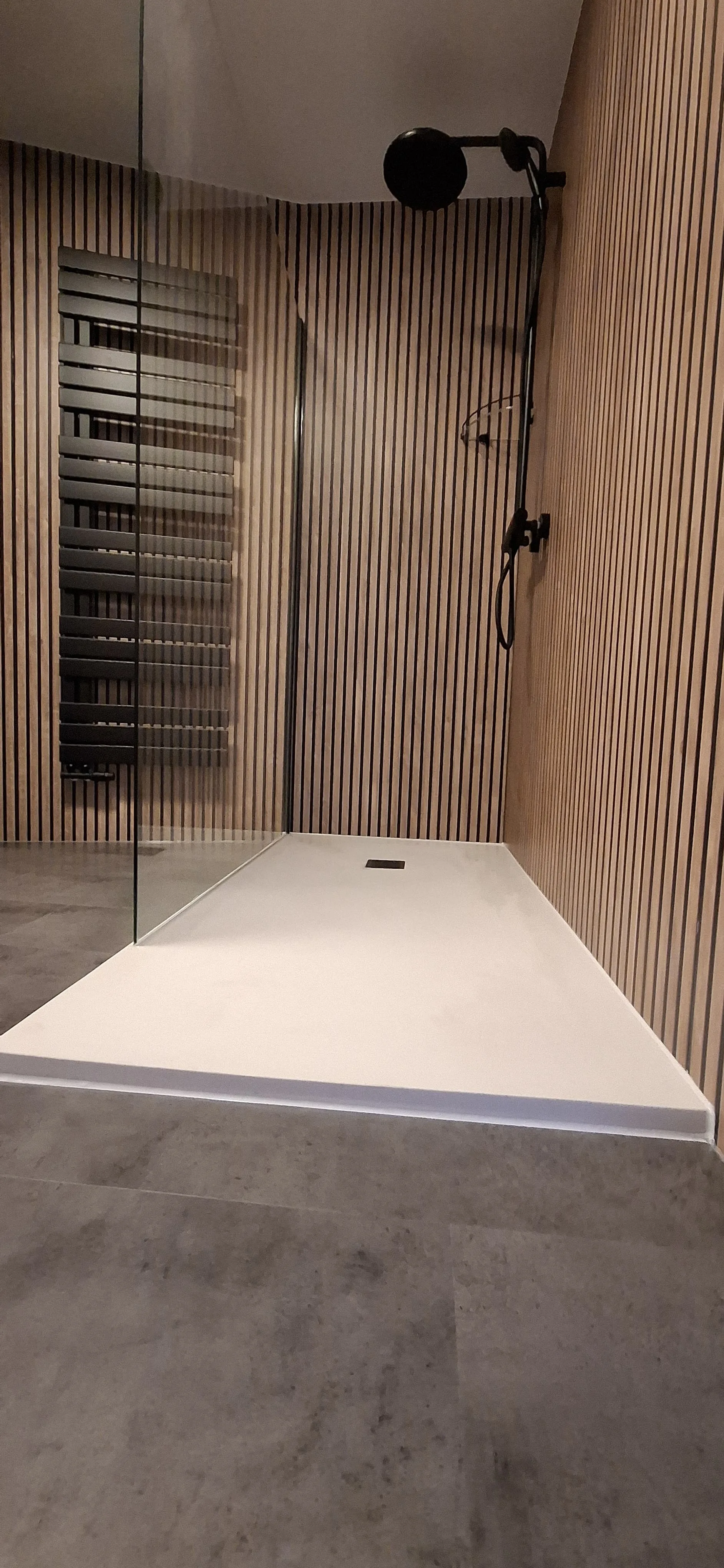 Een moderne douche ruimte met een witte douchebak, een glazen deur, een zwarte douchekop en een wand van verticale houten latten. De vloer is van grijze tegel.