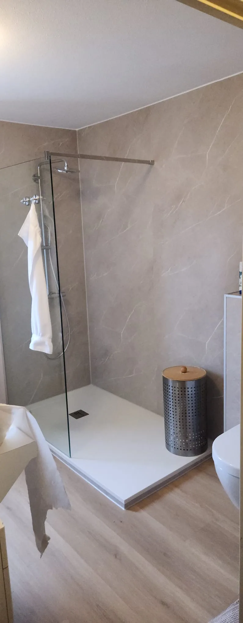 Een moderne badkamer met een glazen douchecabine, betegelde wanden in grijze marmerlook, een witte douchebak en een houten vloer. Er hangt een witte handdoek in de douche.