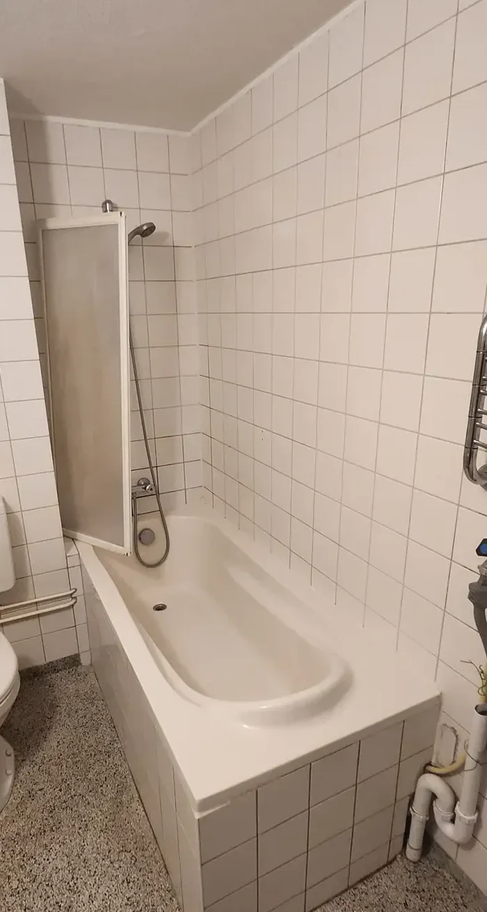 Oude badkamer met ligbad met badscherm, staand toilet met reservoir, witte tegels en spikkelvloer