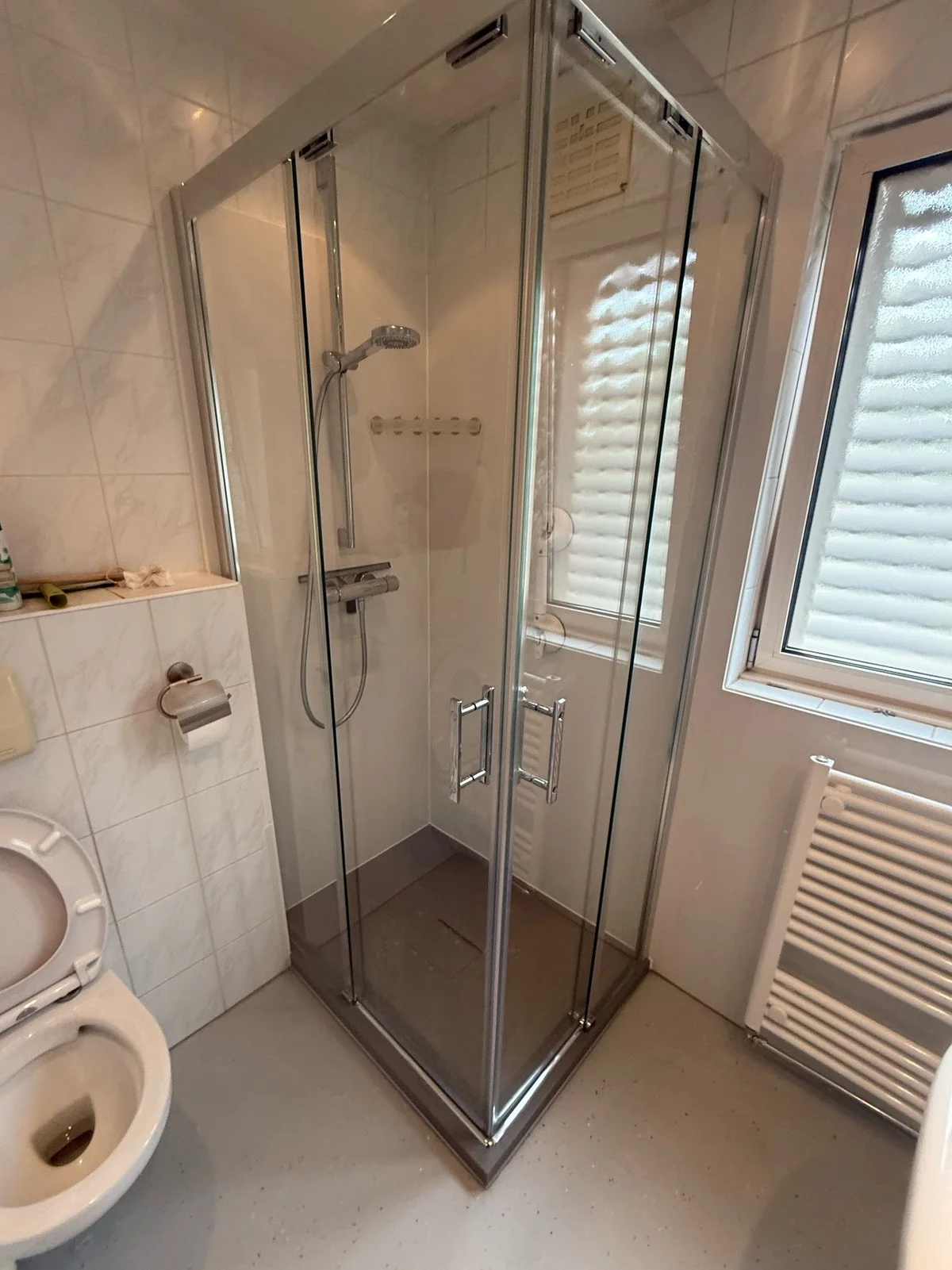 Vierkante glazen douchecabine naast staand toilet en handdoekradiator, nog zonder steunbeugel