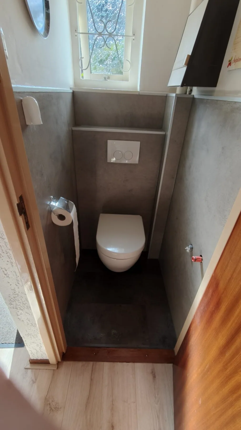 Toilet na renovatie met hangtoilet Gerenoveerd toilet met hangtoilet, grijze betonlook wandpanelen, voorzetwand met bedieningsplaat en raampje met sierrooster