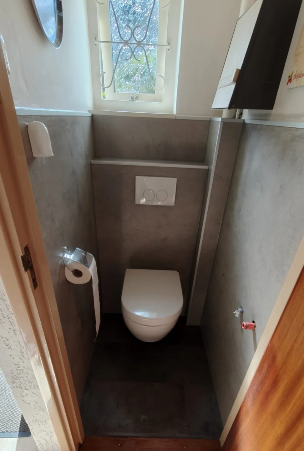 Gerenoveerd toilet met hangtoilet, grijze betonlook wandpanelen, voorzetwand met bedieningsplaat en raampje met sierrooster
